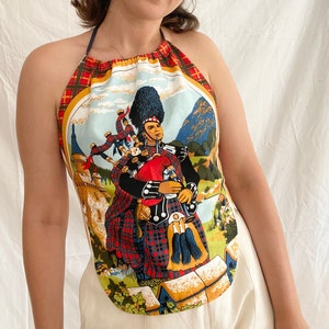 May include: A halter top with a colourful print of a man playing a bagpipe in a Scottish Highlands setting. The print features a castle, mountains, and a plaid kilt.