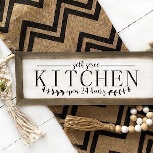May include: A rustic wooden sign with the text "self serve KITCHEN open 24 hours" in black lettering. The sign is surrounded by a hessian runner with black chevron pattern, a macrame plant hanger, and a beaded garland.