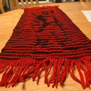 May include: Red and black knitted table runner with fringe. The runner has a geometric pattern.