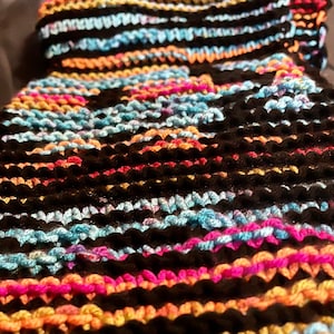 May include: Close-up of a multi-colored knitted fabric with black, orange, pink, and blue yarn. The fabric is woven in a striped pattern.