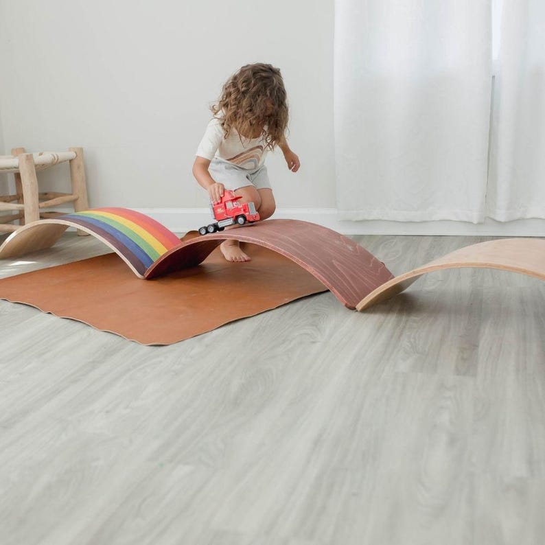Kid playing with a wooden wobble board, using it as a bridge for toy cars while engaging in imaginative pretend play and building balance skills.