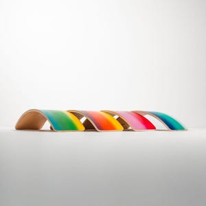 May include: Five rainbow-colored wooden balance boards arranged in a row. Each board features a gradient of vibrant colors, transitioning smoothly from one hue to the next. The colors include teal, yellow, orange, pink, and a deeper blue.  These wooden balance boards are designed for children's play and physical development, promoting balance and coordination skills. The smooth, curved shape of the boards is clearly visible, along with the high-quality finish of the paint.