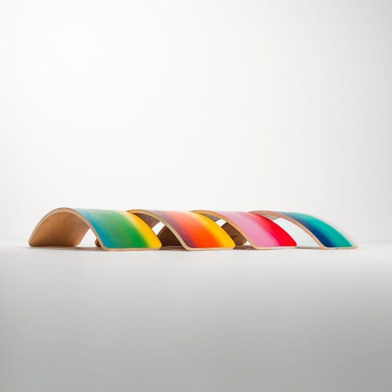 May include: Five rainbow-colored wooden balance boards arranged in a row. Each board features a gradient of vibrant colors, transitioning smoothly from one hue to the next. The colors include teal, yellow, orange, pink, and a deeper blue.  These wooden balance boards are designed for children's play and physical development, promoting balance and coordination skills. The smooth, curved shape of the boards is clearly visible, along with the high-quality finish of the paint.