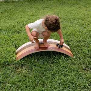 Kid sitting outdoors on a wooden wobble board, using it as a track for toy cars while enjoying active play and developing coordination.