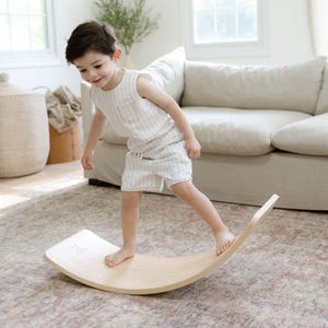 A young child is using the balance board in a cozy playroom, which is a great tool for developing balance, coordination, and core strength. The board is made of natural wood and has a simple, minimalist design. It is suitable for indoor use and is a fun way for children to improve their motor skills.