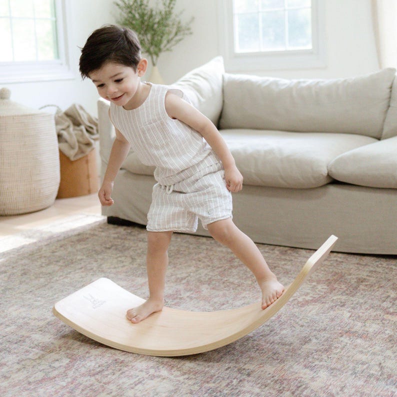 A young child is using the balance board in a cozy playroom, which is a great tool for developing balance, coordination, and core strength. The board is made of natural wood and has a simple, minimalist design. It is suitable for indoor use and is a fun way for children to improve their motor skills.