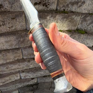 May include: A vintage hunting knife with a curved, polished steel blade. The handle is wrapped in dark leather with brown and red accents. The knife has a metal bolster and pommel.