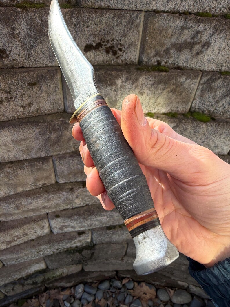 May include: A vintage hunting knife with a curved, polished steel blade. The handle is wrapped in dark leather with brown and red accents. The knife has a metal bolster and pommel.