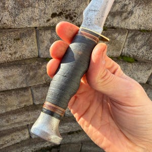 May include: A vintage hunting knife with a worn, textured blade and a dark leather-wrapped handle. The knife features a brass bolster and a metal pommel. The blade shows signs of use, suggesting it's a well-loved tool.