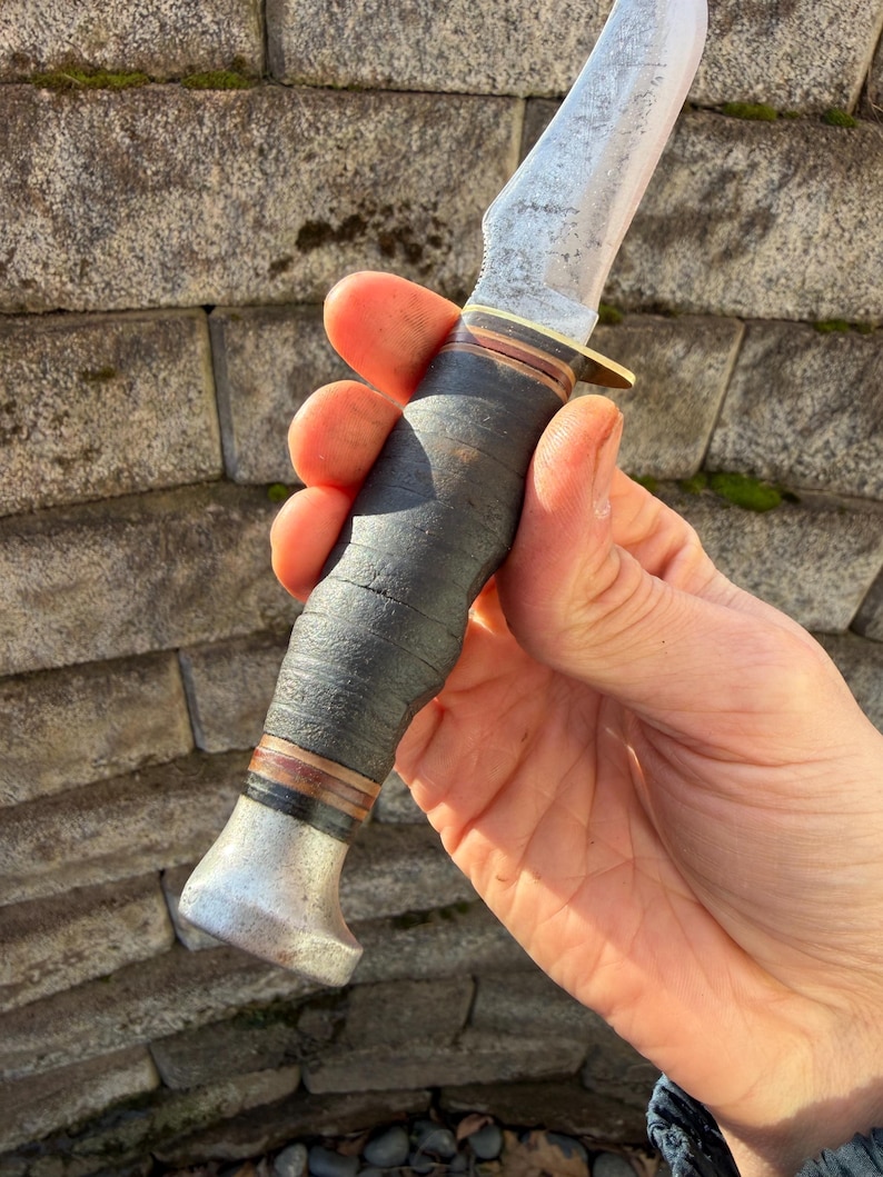 May include: A vintage hunting knife with a worn, textured blade and a dark leather-wrapped handle. The knife features a brass bolster and a metal pommel. The blade shows signs of use, suggesting it's a well-loved tool.