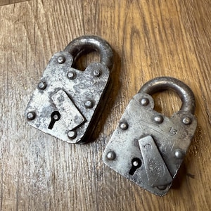 May include: Two antique metal padlocks with a weathered, aged appearance. Each lock has a curved handle and a rectangular body with rivets. One lock has the number 13 stamped on it.