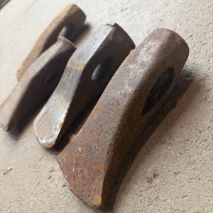May include: Four weathered, rusty metal splitting mauls are arranged on a concrete surface. The mauls vary in size and shape, with a wedge-like design and a hole for a handle. The tools show signs of age and use, with a brown and orange color.