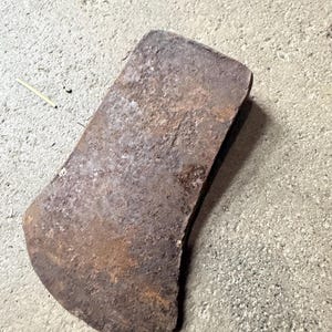 May include: A weathered, rusty axe head with a curved blade. The metal has a rough, textured surface with areas of dark brown and orange discoloration, suggesting age and exposure to the elements. The axe head is lying on a concrete surface.