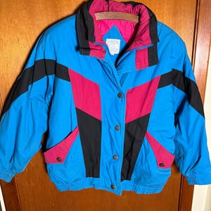 May include: A vintage blue, black, and pink jacket with a geometric design. The jacket has a high collar, button closure, and two front pockets. The interior lining is pink. The jacket is hanging on a metal hanger.