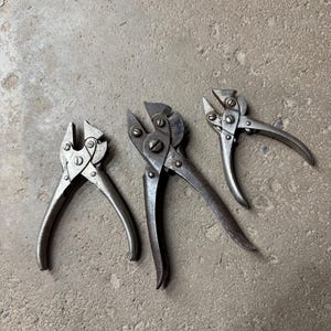May include: Three vintage metal pliers with curved handles are arranged on a concrete surface. The pliers have a silver finish and show signs of wear, suggesting they are antique tools. The jaws of the pliers are open, ready for use.