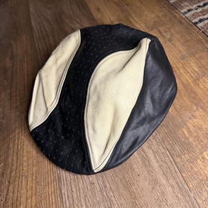 May include: A black and cream-colored newsboy cap. The cap features a perforated black section and cream-colored panels. The cap is made of leather and has a classic design.