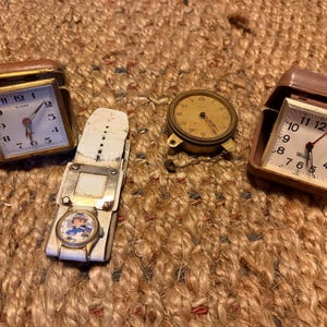 May include: A collection of four timepieces including two square travel alarm clocks in brown cases, one round brass pocket watch with a small chain, and one wristwatch with a white leather strap featuring a circular graphic of a cartoon character.