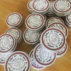 May include: A close-up of multiple small, round, silver tins with red and black labels. The labels feature a stylized illustration of a fox holding an axe and the text "DEFOREST AXE AND TOOLWORKS Classic Wax".