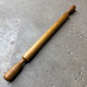 May include: A wooden rolling pin with a light brown finish. The rolling pin has a cylindrical body and two rounded handles. The wood grain is visible, and the pin is resting on a gray surface.