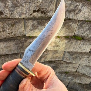 May include: A handcrafted knife with a light-colored blade and a dark handle. The blade has a slightly curved edge and a textured surface. The handle is wrapped in dark leather with brass and brown accents. The knife is held against a stone wall.