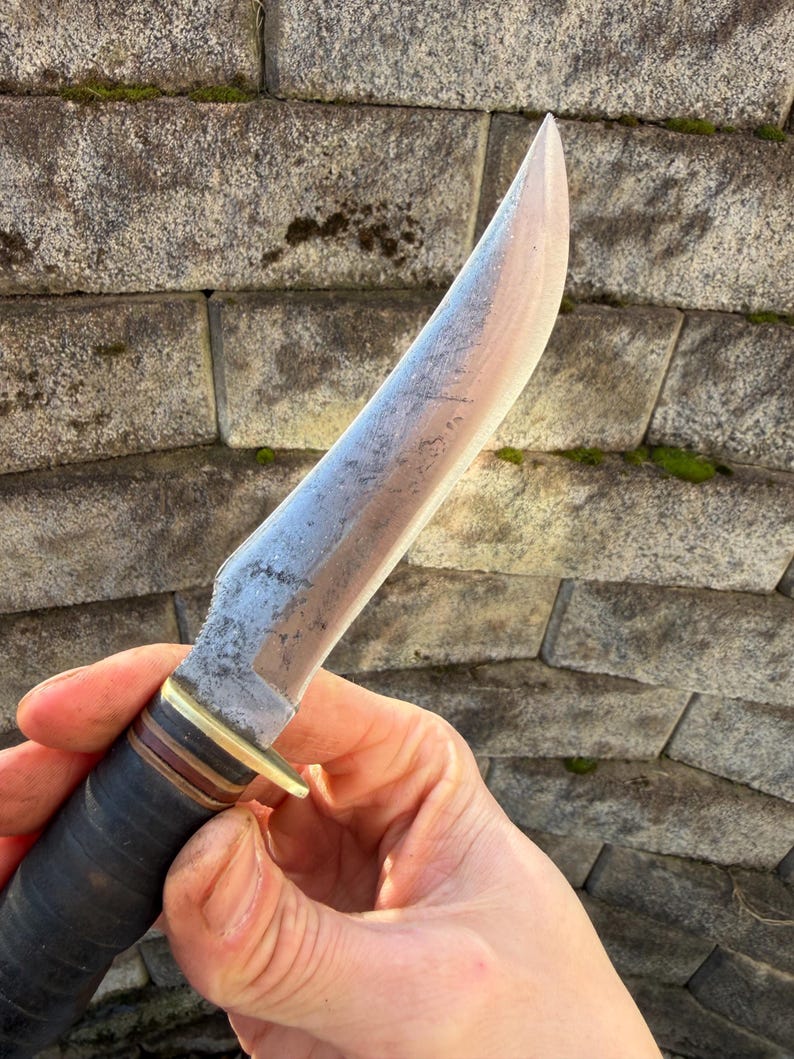 May include: A handcrafted knife with a light-colored blade and a dark handle. The blade has a slightly curved edge and a textured surface. The handle is wrapped in dark leather with brass and brown accents. The knife is held against a stone wall.