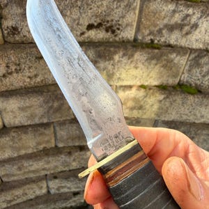 May include: A vintage Ka-Bar knife with a curved blade. The knife features a leather handle and a brass guard. The blade shows signs of wear, and the text "Ka-Bar 1232 USA" is visible on the blade.
