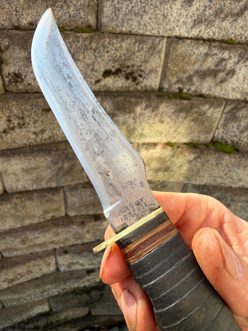 May include: A vintage Ka-Bar knife with a curved blade. The knife features a leather handle and a brass guard. The blade shows signs of wear, and the text "Ka-Bar 1232 USA" is visible on the blade.