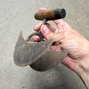 May include: A vintage, double-bladed mezzaluna knife with a wooden handle. The blades are curved and made of aged metal, showing signs of wear. The handle is a dark brown color.