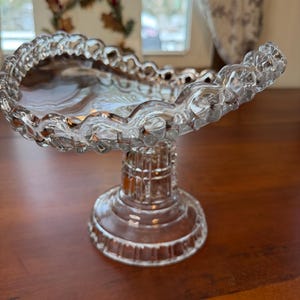 May include: A clear, decorative glass cake stand with a scalloped edge. The stand has a round base and a detailed stem. The glass has a textured, cut-glass appearance, reflecting light. The stand is on a wooden surface.