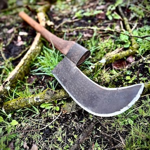 May include: A vintage-style curved blade axe with a wooden handle, resting on a bed of green grass and twigs. The axe head is dark with a sharp, curved edge. The handle is a light brown color.