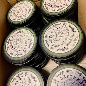 May include: A close-up of several black and green containers of axe wax. The containers are stacked in a cardboard box. The labels on the containers read "DEFOREST AXE AND TOOLWORKS Black Pine Axe Wax".