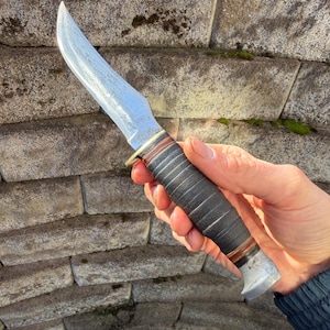 May include: A vintage hunting knife with a curved blade. The blade has a textured surface. The handle is black with a brown and silver accent. The knife is held against a stone wall.