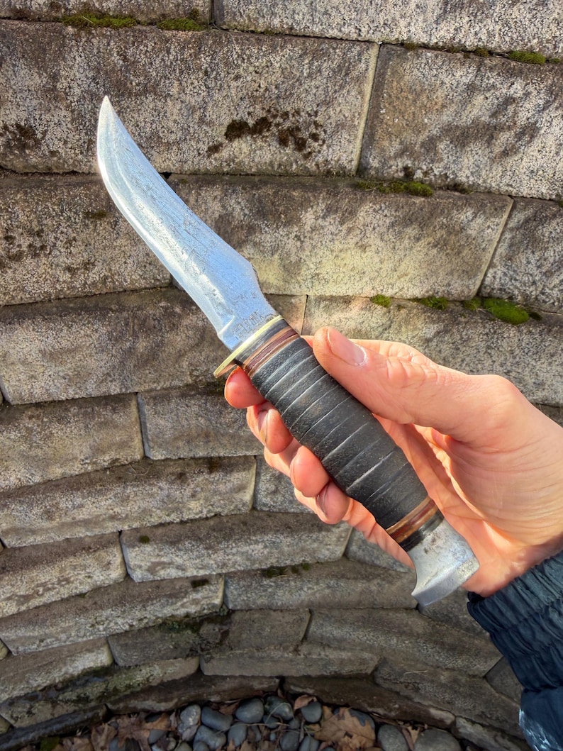 May include: A vintage hunting knife with a curved blade. The blade has a textured surface. The handle is black with a brown and silver accent. The knife is held against a stone wall.