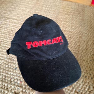 May include: A black baseball cap featuring the word TOMCATS embroidered in red thread on the front panel. The cap has a structured crown and a curved brim, with a visible fabric texture and a small black strap visible on the left side.