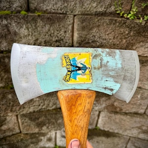 May include: A vintage double-bit axe with a wooden handle and a weathered metal head. The axe head has a faded blue paint and a "FOREST KING" sticker. The axe is held against a textured stone wall.