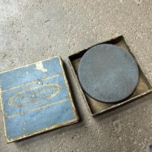 May include: A Craftsman brand sharpening stone in its original box. The box is blue with gold lettering and a gold border. The stone is round and gray, and sits inside a brass-colored metal container.