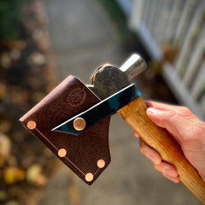 May include: A small axe with a wooden handle and a metal head, secured by a black and teal accent. The axe is partially covered by a brown leather sheath with copper rivets. The sheath has a logo embossed on it.
