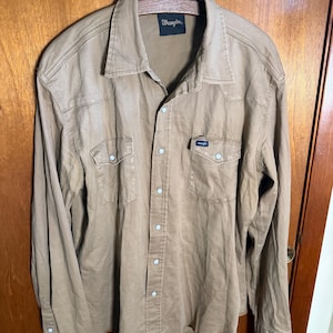 May include: A tan Wrangler button-down shirt with long sleeves, two chest pockets, and pearl snap closures. The shirt has the Wrangler logo on the chest pocket and the collar. The shirt is hanging on a metal hanger.
