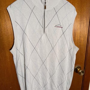 May include: A light beige sleeveless sweater vest with a diamond pattern and a mock neck. The vest has a quarter-zip closure and the word "LEDGEROCK" embroidered in red on the chest.