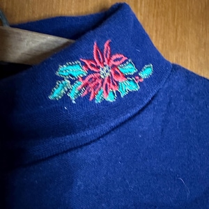 May include: Close-up of a navy blue turtleneck sweater. The collar features an embroidered poinsettia design with red petals and green leaves. The sweater is made of a soft, possibly knitted, material.