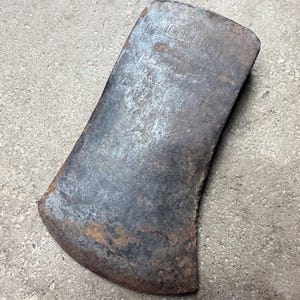 May include: A vintage axe head with a weathered, rusty appearance. The metal tool has a curved blade and the word "COLLINS" is stamped on the top. The axe head is set against a gray concrete surface.