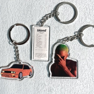 Op de afbeelding: Drie sleutelhangers met de albumhoes van Frank Oceans Blonde. Eén sleutelhanger is een rode auto, een andere is een zwart-witte afbeelding van Frank Ocean en de derde is een witte rechthoek met de albumtitel en de tracklist.