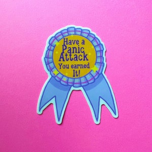 May include: A blue and yellow ribbon with the text "Have a Panic Attack You earned It!" printed on it. The ribbon has a star design and is on a pink background.