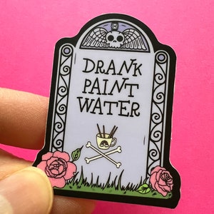 May include: A sticker of a tombstone with the text "DRANK PAINT WATER" and a skull and crossbones with a cup of paintbrushes. The tombstone is surrounded by green grass and pink roses.