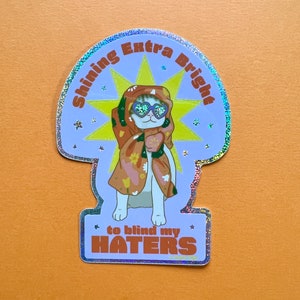 May include: A holographic sticker featuring a cartoon cat wearing sunglasses and a patterned robe. The cat is surrounded by a yellow star with the text "Shining Extra Bright" above the cat and "to blind my HATERS" below the cat.