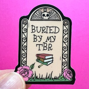 May include: A black and white sticker of a tombstone with the text "BURIED BY MY TBR" and a stack of books on top. The tombstone has a skull and wings at the top and a decorative border.