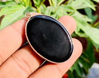 May include: Oval-shaped black pendant with a gold-colored metal border. The pendant is smooth and reflective, with a polished surface. The jewelry is held against a backdrop of green leaves and a red pot.