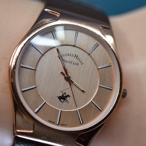 May include: A gold-toned wristwatch with a white face and black hands. The watch face has the words "Beverly Hills Polo Club" and "Japan Movt" printed on it.