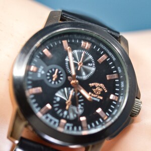May include: A black chronograph wristwatch with a black leather band. The watch face has a black background with rose gold accents and a tachymeter scale. The watch face also features the words "TACHYMETRE" and "U.S. POLO ASSN."