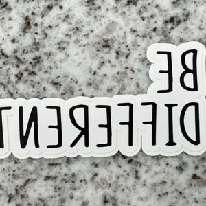 May include: White sticker with black text that reads "BE DIFFERENT" on a gray speckled background.